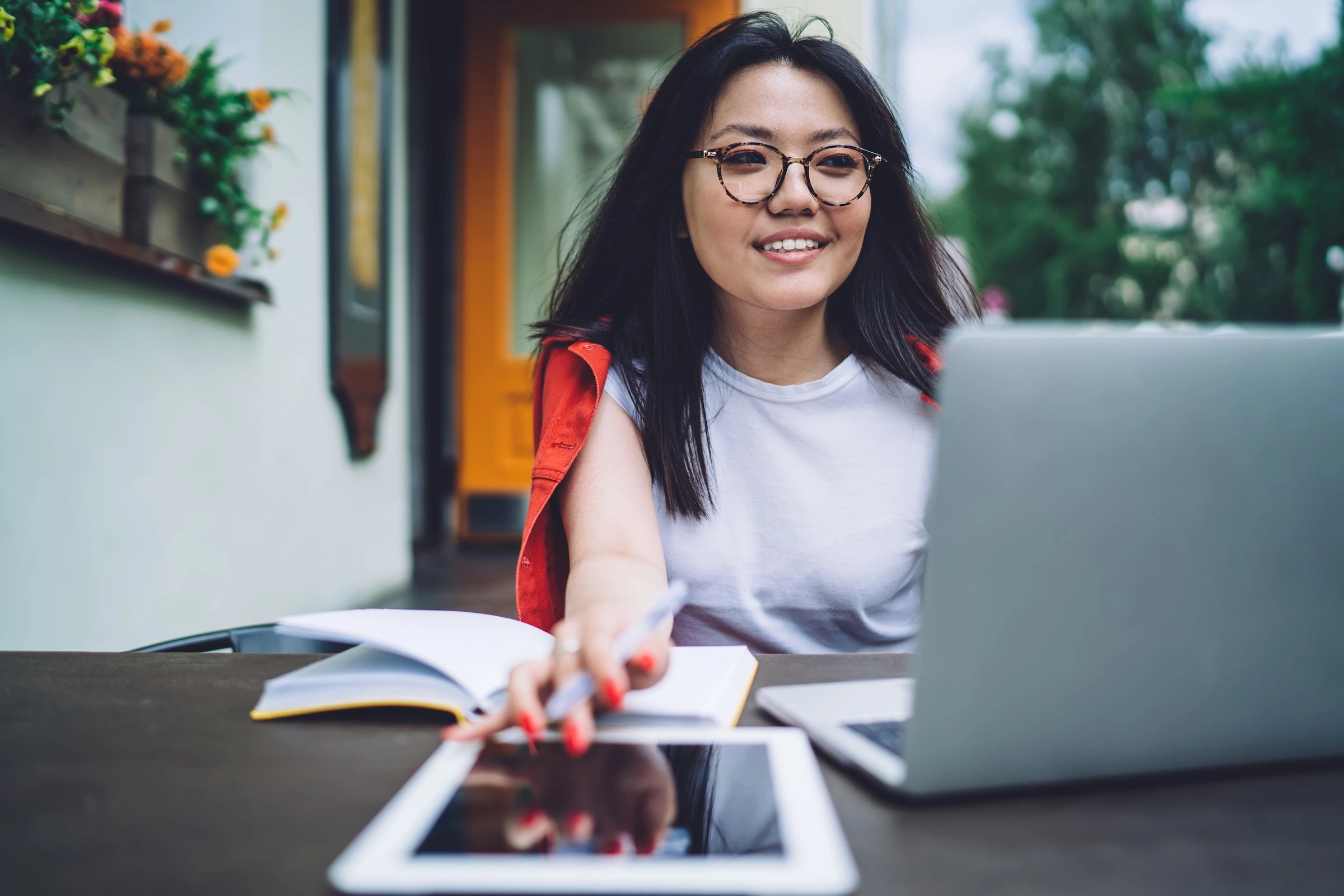 A smiling young woman with long black hair and glasses works on a laptop at an outdoor cafe table. She is reaching for a tablet.