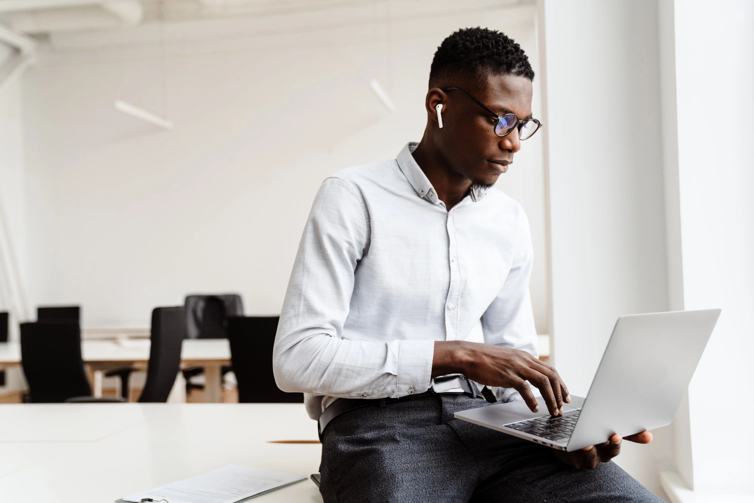 A man in glasses sits on a desk in a modern office, focused on his laptop. Black office chairs are visible in the blurred background.