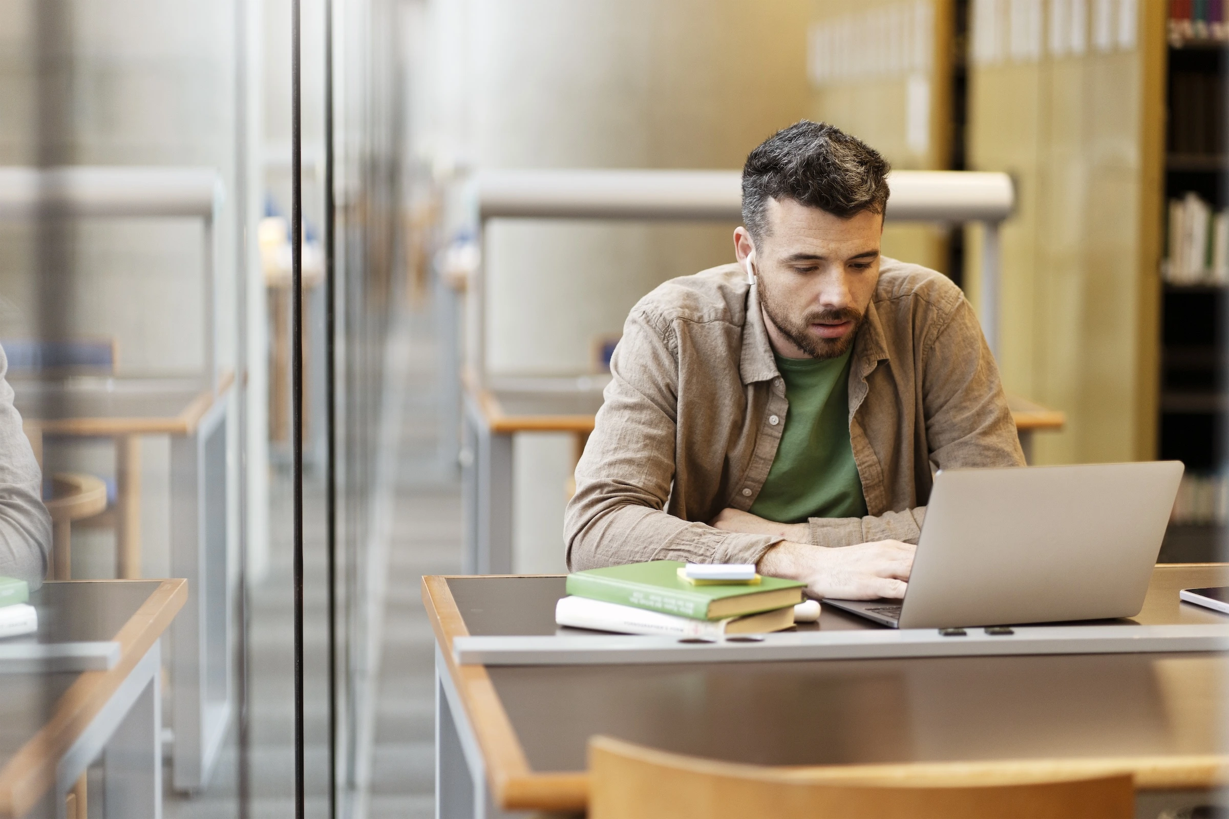 A man in a library works on a laptop with green books stacked nearby. The background shows empty study carrels and bookshelves.