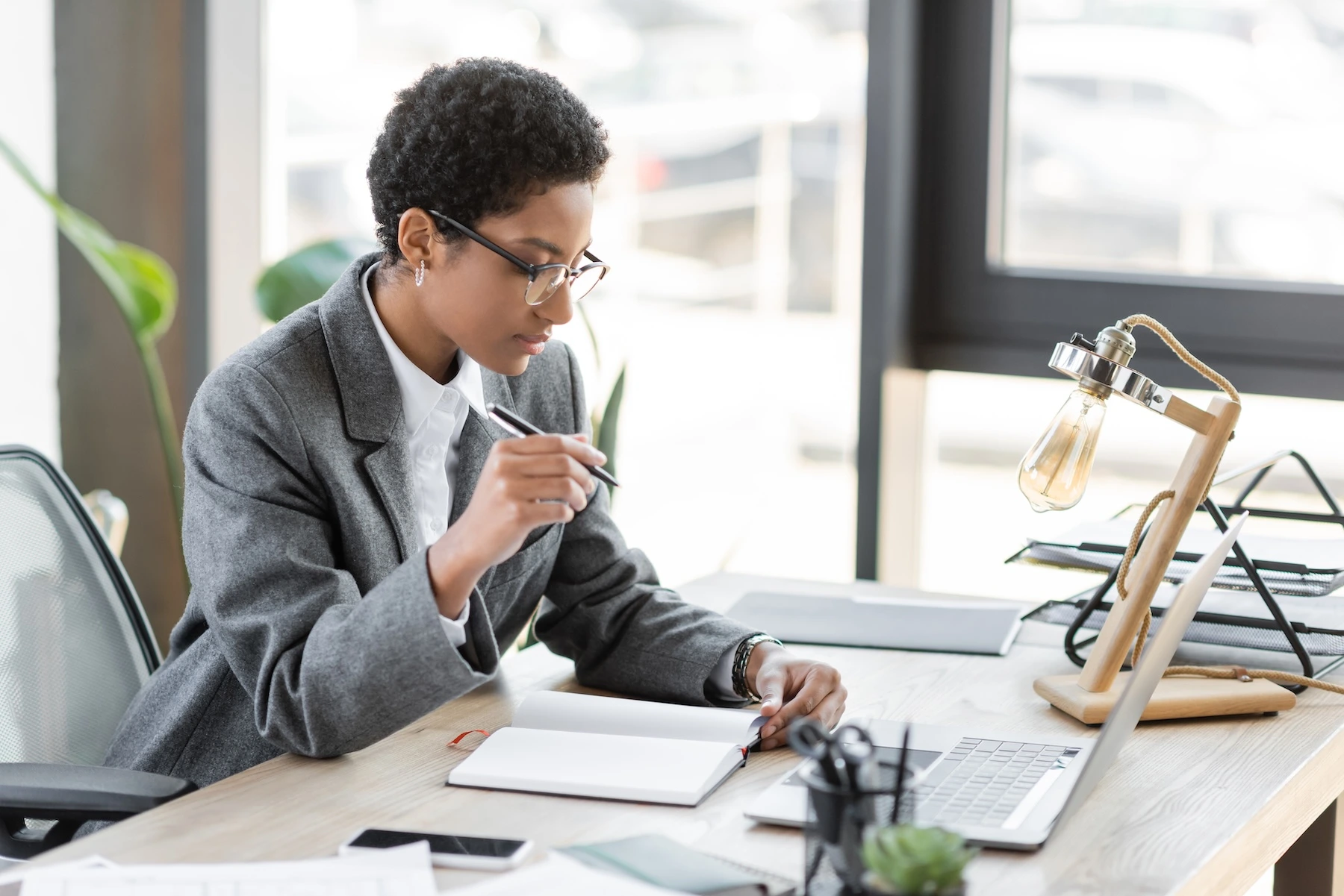 "A professional woman in a grey blazer taking notes at a desk while looking at a laptop in a bright, modern office."