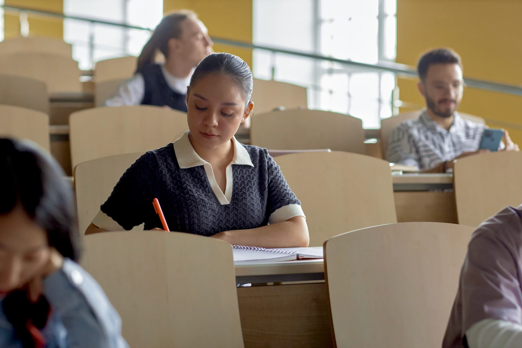 A female student sits at a desk in a lecture hall, focused on writing in her notebook while other students sit nearby.
