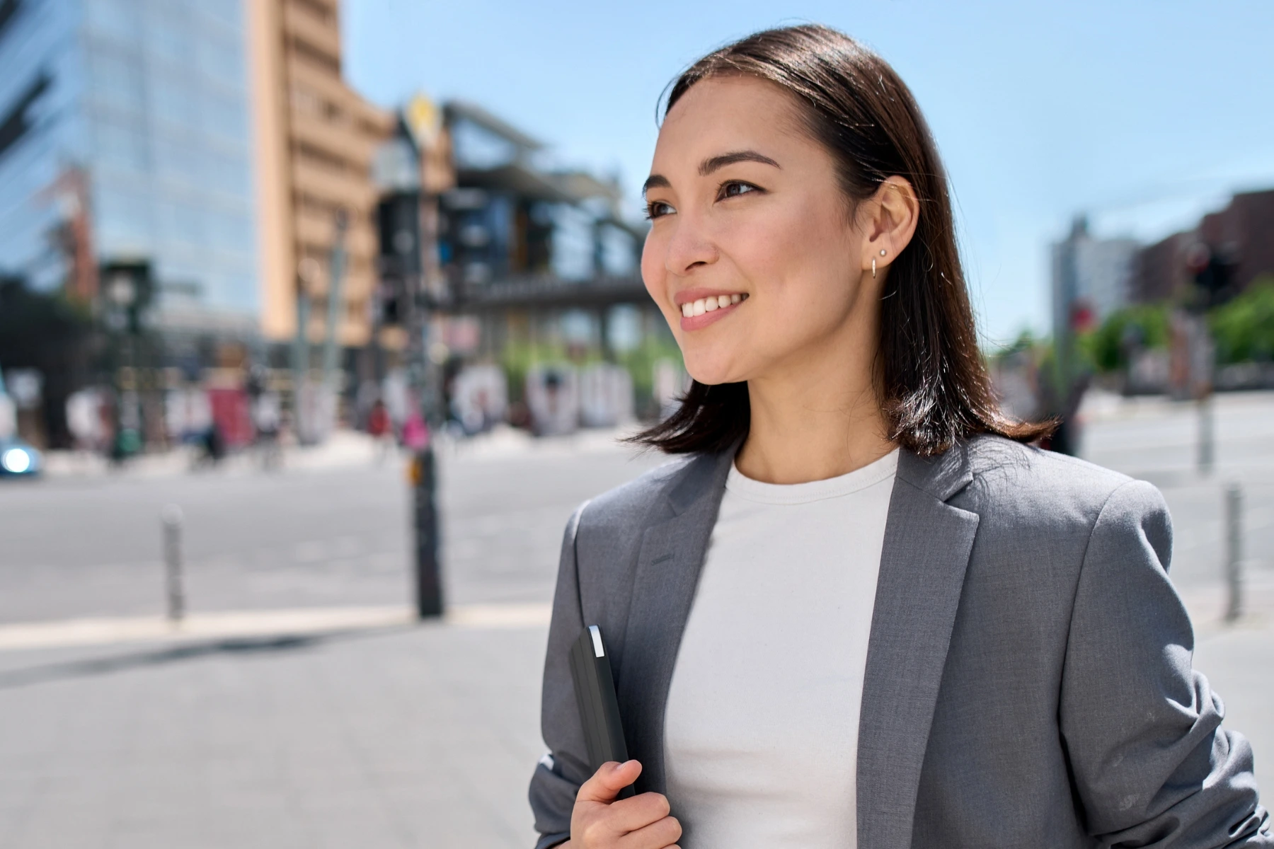 A smiling woman in a grey blazer walks down a sunny city street holding a laptop or tablet under her arm.