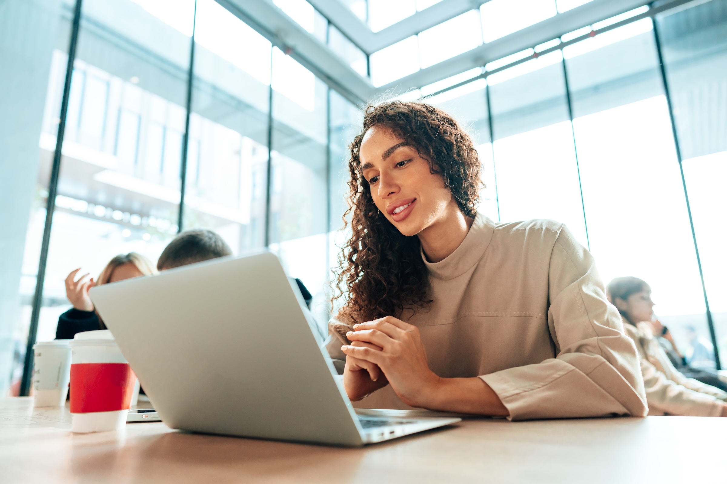 A woman with curly hair smiles while working on a laptop in a bright, modern building with large glass windows.
