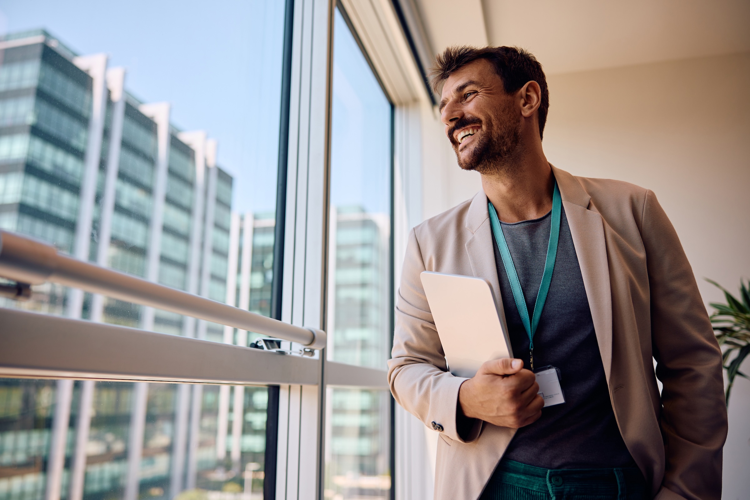A smiling businessman in a blazer holds a tablet and looks out a large window at a city office building.