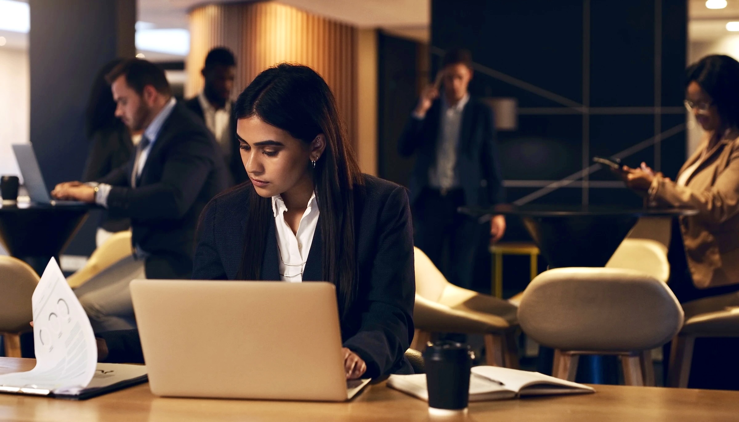 A businesswoman in a suit focuses on her laptop at a table in a busy, modern office lounge.