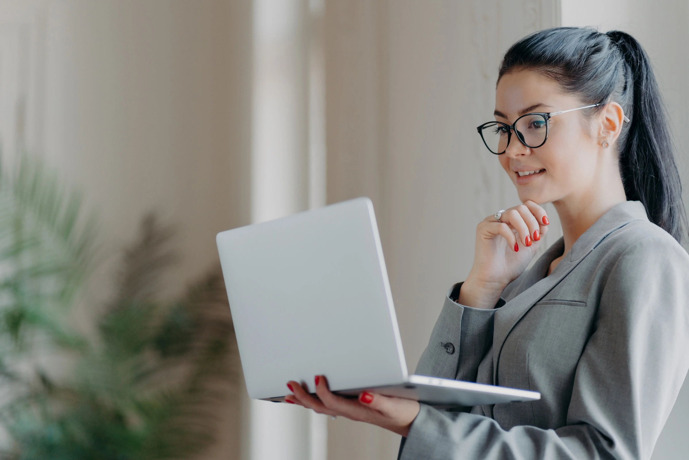 A smiling woman with glasses and dark hair in a ponytail holds an open laptop while looking at the screen.